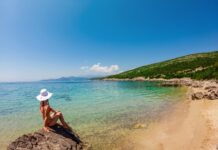Istanbul Beaches in 2026: Where to Swim Safely Woman in a hat sitting on rocks by clear blue Turkish coast water, framed by mountains