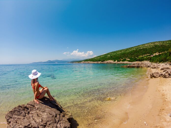 Woman in a hat sitting on rocks by clear blue Turkish coast water, framed by mountains